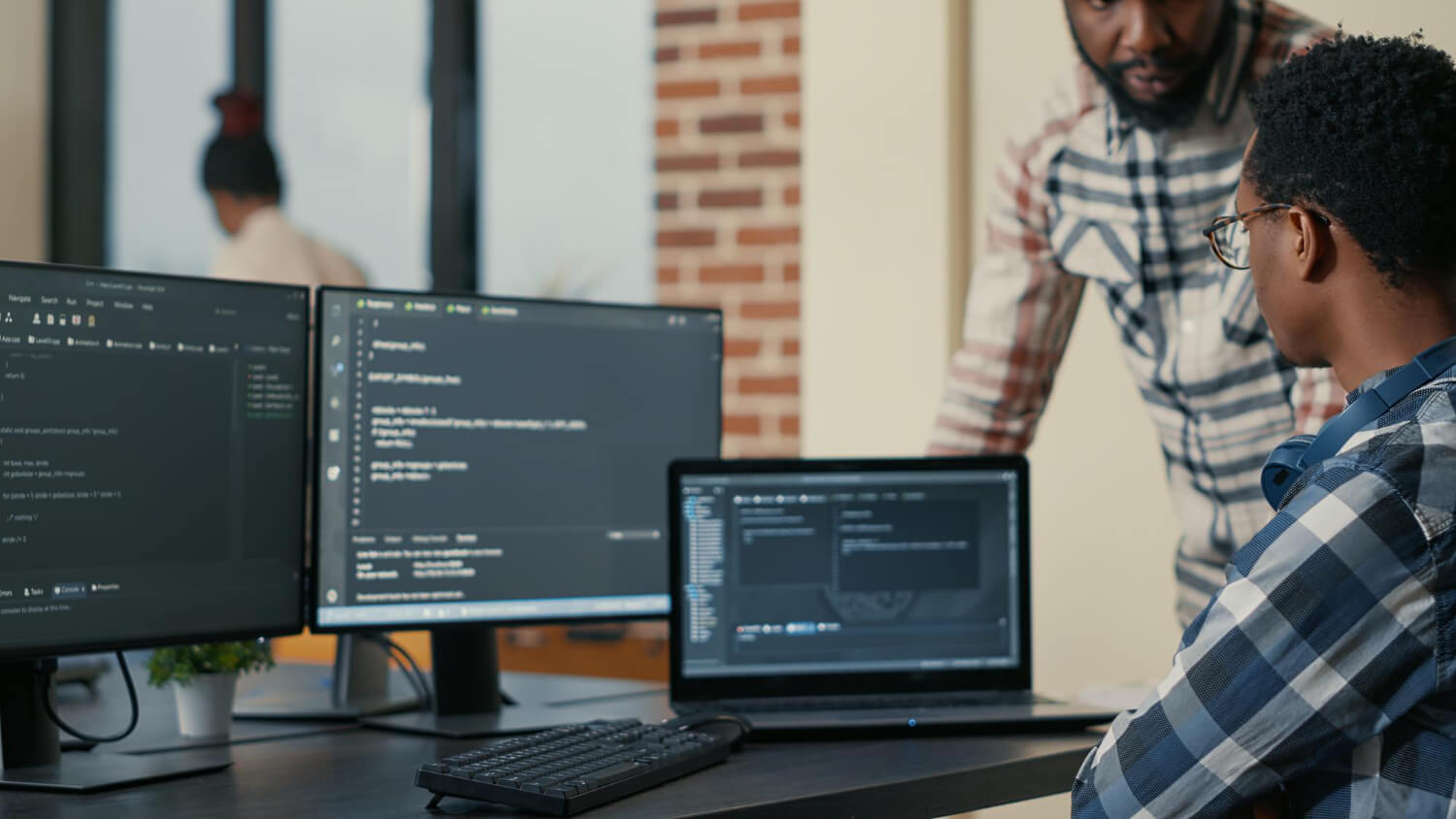 Dos personas en oficina trabajando en tres pantallas con codigo