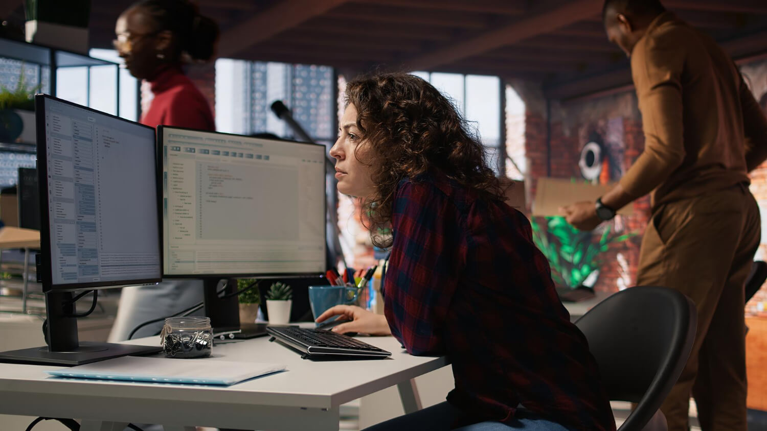 Persona concentrada programando en oficina con monitores duales. Dos personas mas al fondo