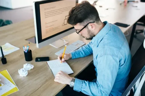 Persona en escritorio escribiendo en cuaderno, con computadora y bolígrafos alrededor