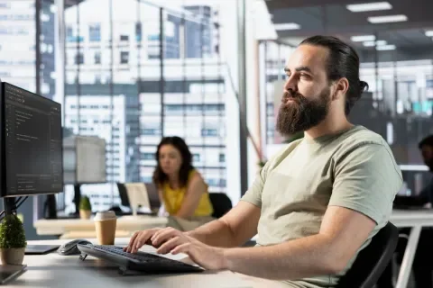 Hombre con barba trabajando en computadora en oficina moderna; fondo con mujer y ventanas grandes