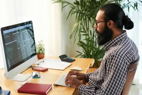 Hombre trabajando en computadora con código, escritorio ordenado, planta y ventana al fondo.